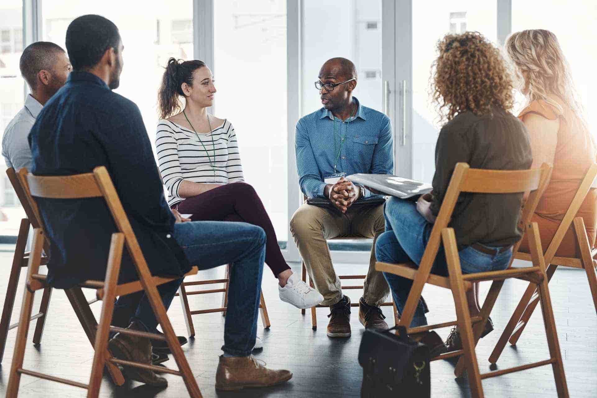 Diverse group of adults seated in a circle participating in a group discussion in a bright treatment-style setting.