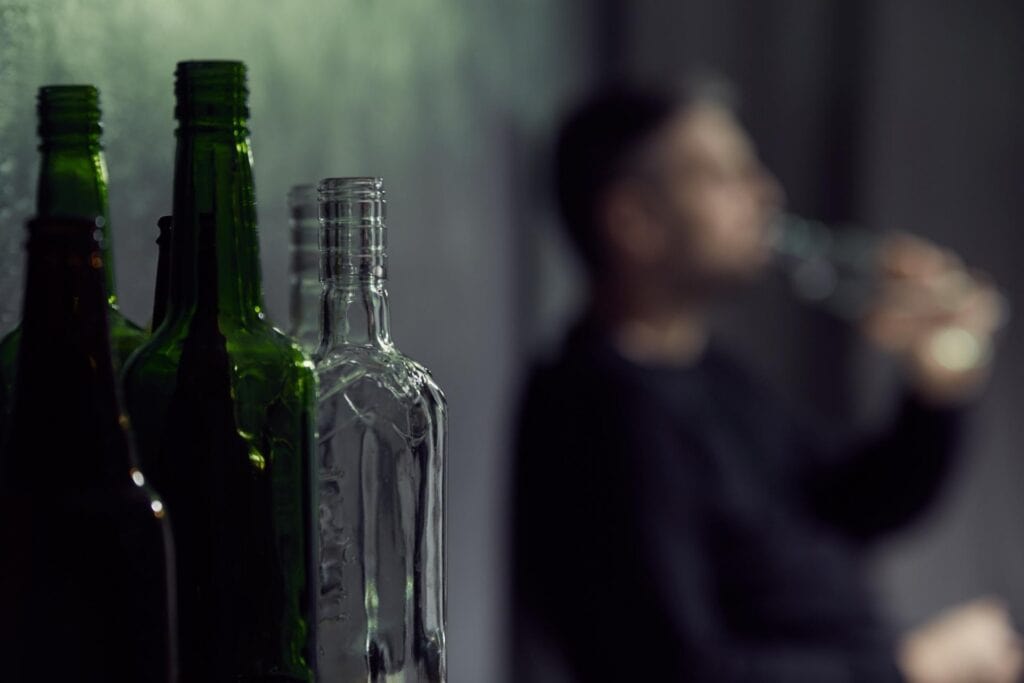 Early Signs Of Alcohol Addiction Most People Miss 1 Empty alcohol bottles in the foreground with a person drinking alone in the background, representing alcohol addiction.